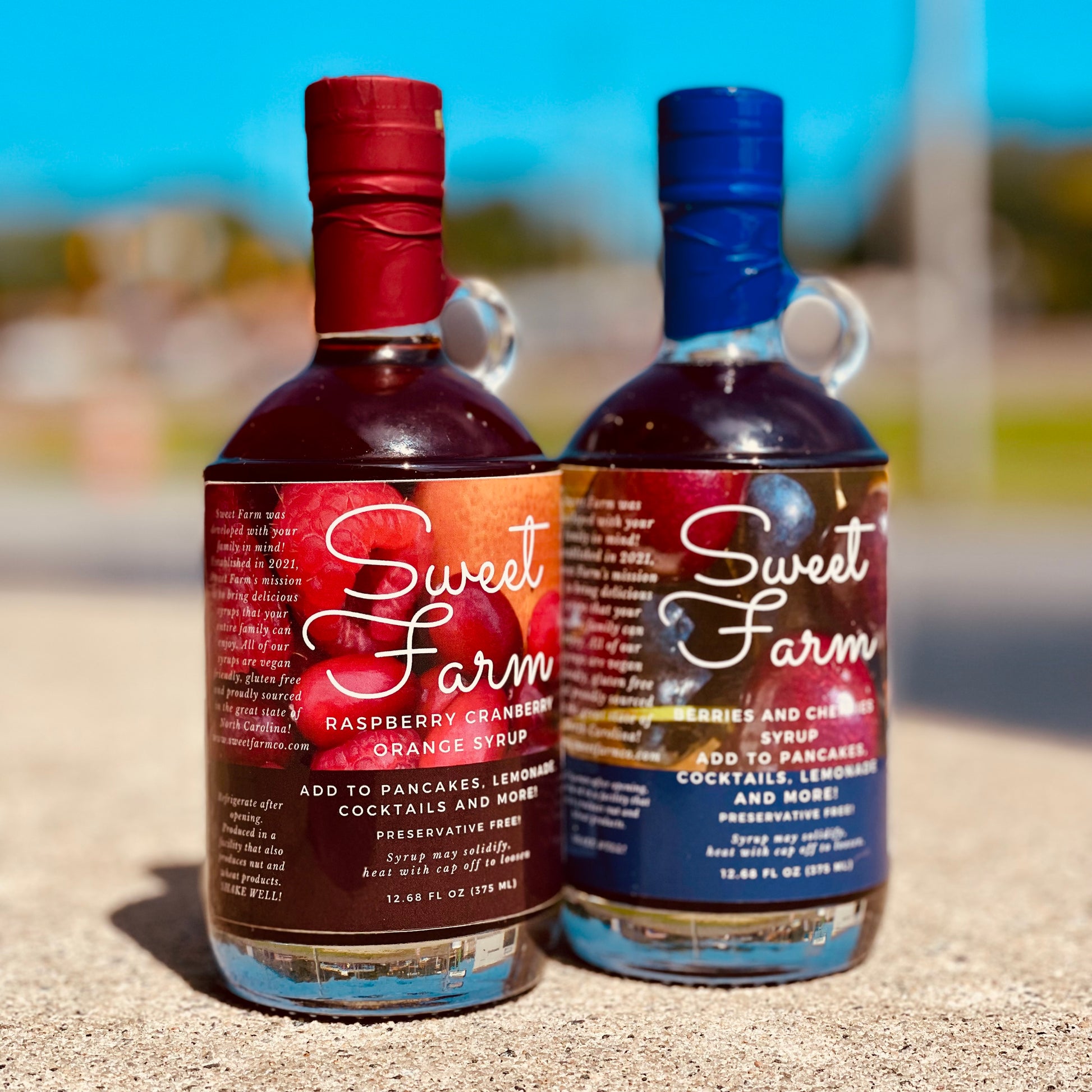 Two bottles of Sweet Farm syrup on a sandy surface with a blurred outdoor background.