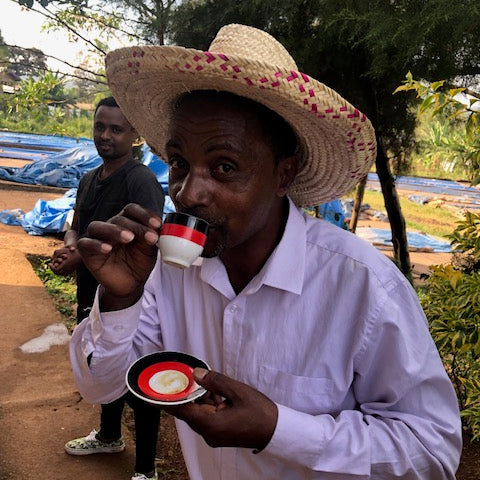 Muetefa Abakeno wearing a straw hat and white shirt, sipping from a demitasse and holding a matching saucer, which is white, red, and black.
