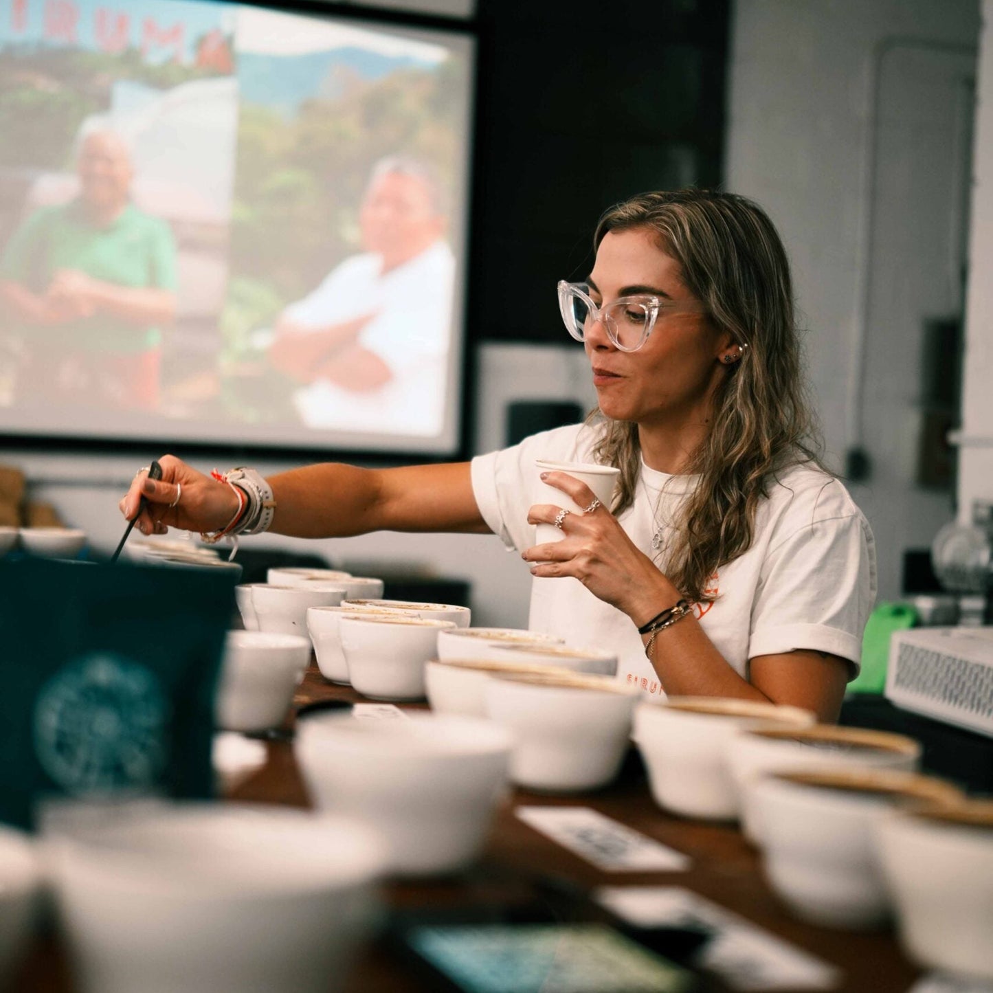 Valentina Duque standing behind a table full of white cupping bowls. She is rinsing her spoon in between sips.